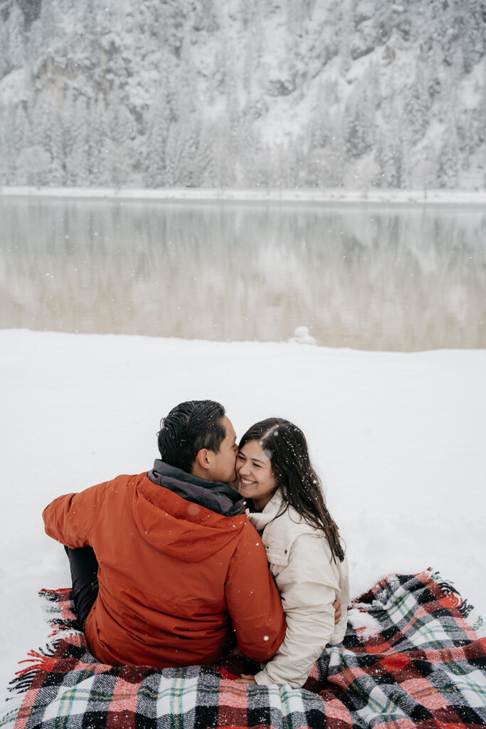 Couple sitting on snowy blanket by a lake.