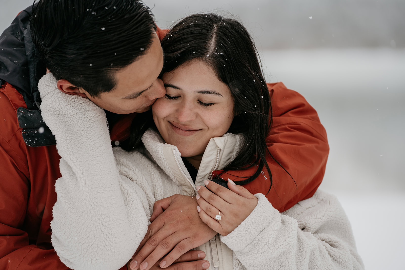 Couple embracing in snowy winter setting, engagement ring visible.