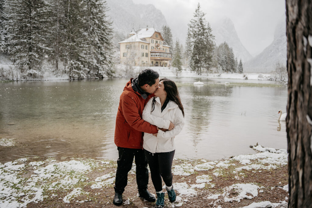 Couple kissing by snowy lake with mountains.