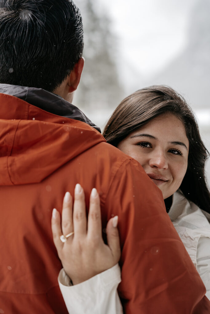 Woman hugging man, wearing engagement ring, snowy background.