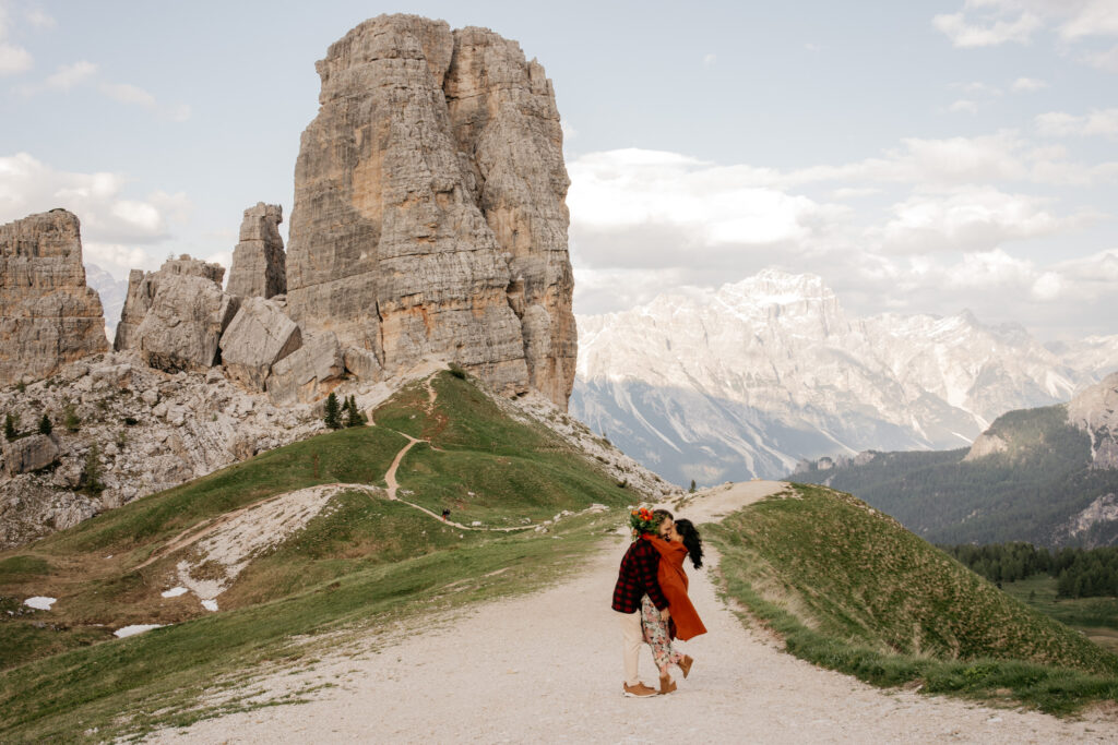 Couple embracing on mountain path, rugged peaks backdrop.