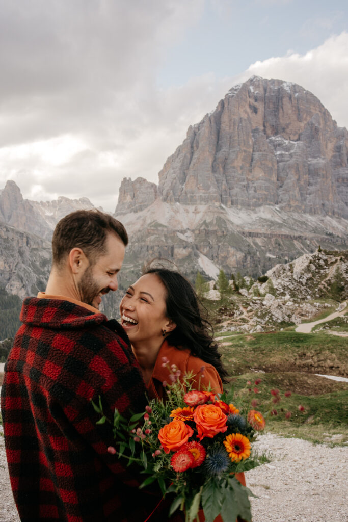 Couple laughing with mountain backdrop, holding flowers.