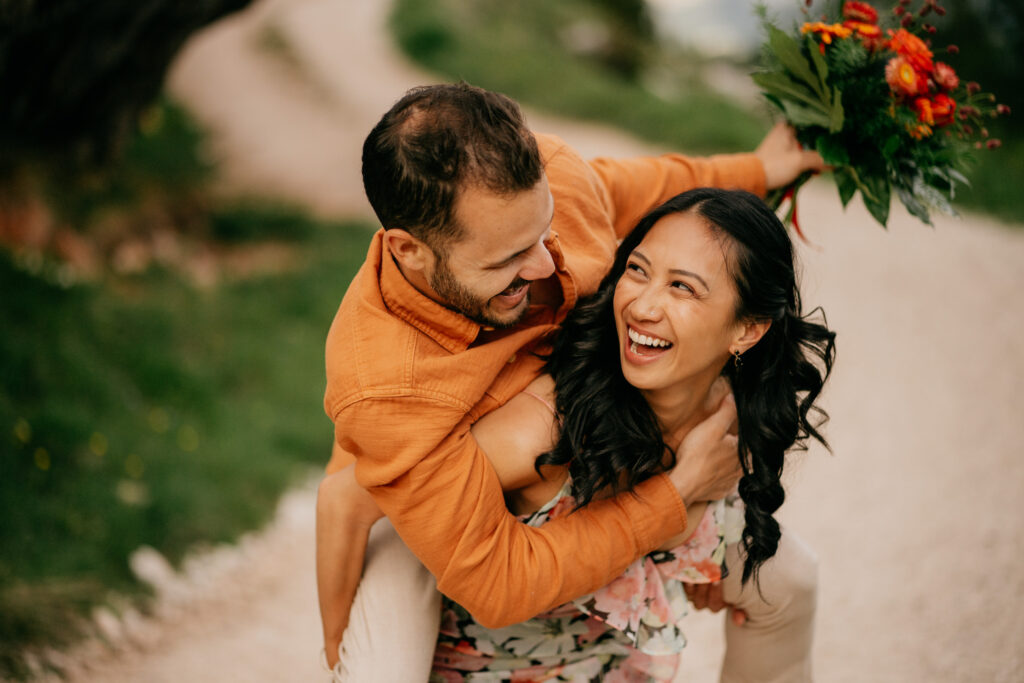 Smiling couple piggyback, holding flowers outdoors