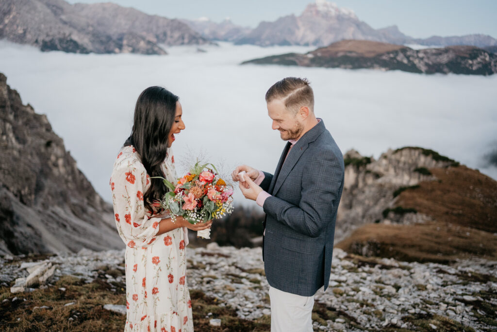 Couple engaged on mountain, holding flowers.