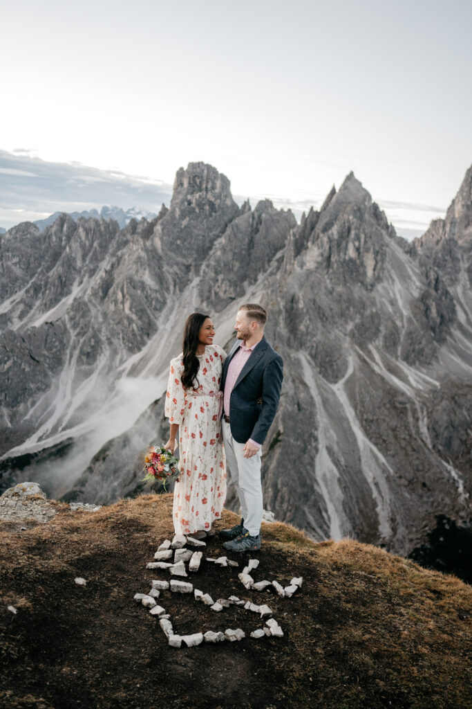 Couple smiling on mountain peak with rocky backdrop.