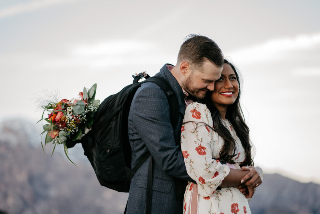 Couple embraces with bouquet, mountains in background.