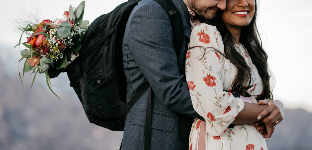 Couple embracing with bouquet against mountain backdrop.