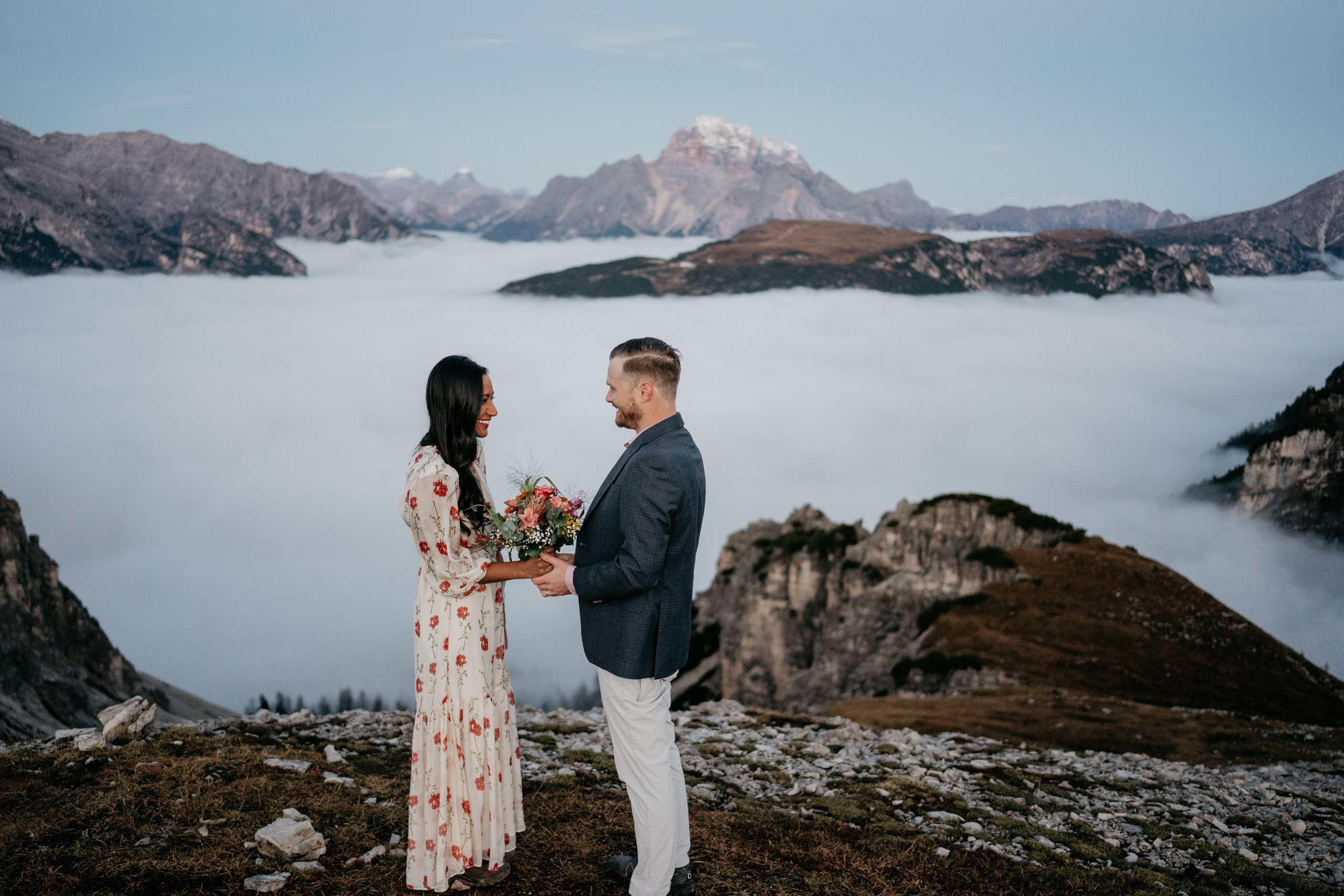 Couple standing on mountain, holding flowers, smiling.