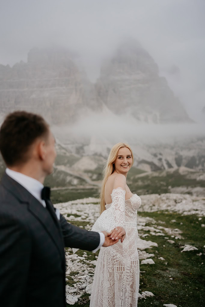 Bride in lace dress holding groom's hand in mountains.