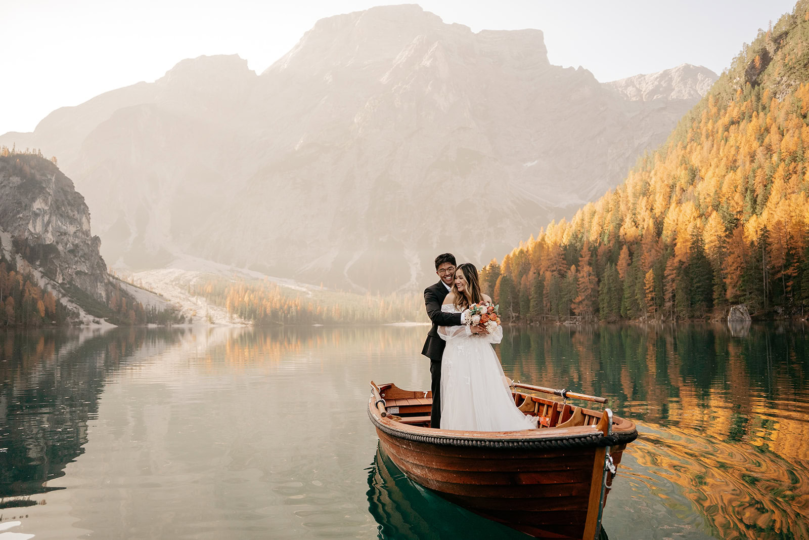 Bride and groom in boat on lake, autumn backdrop.