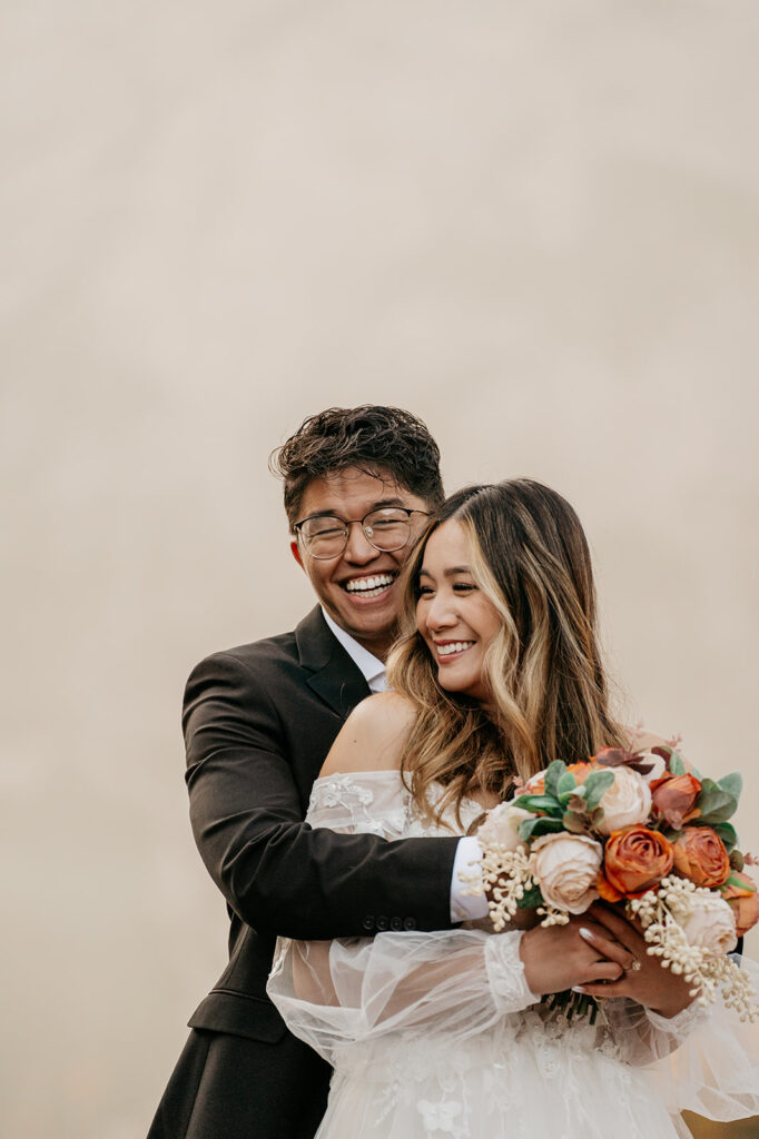 Smiling couple embraces in wedding attire with flowers.