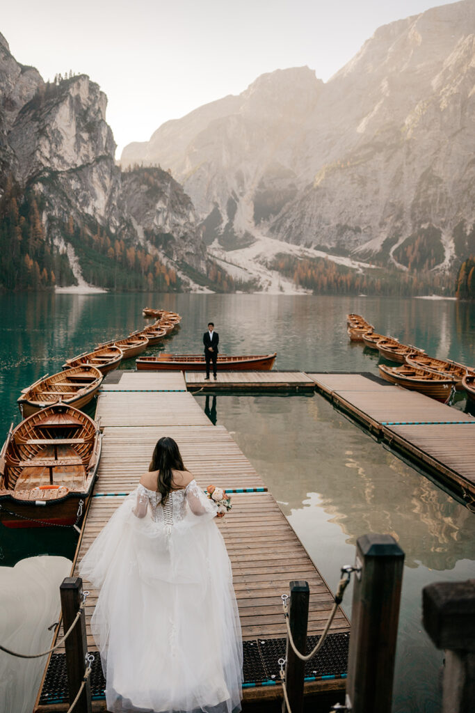 Bride approaches groom on lake dock, scenic mountain backdrop.