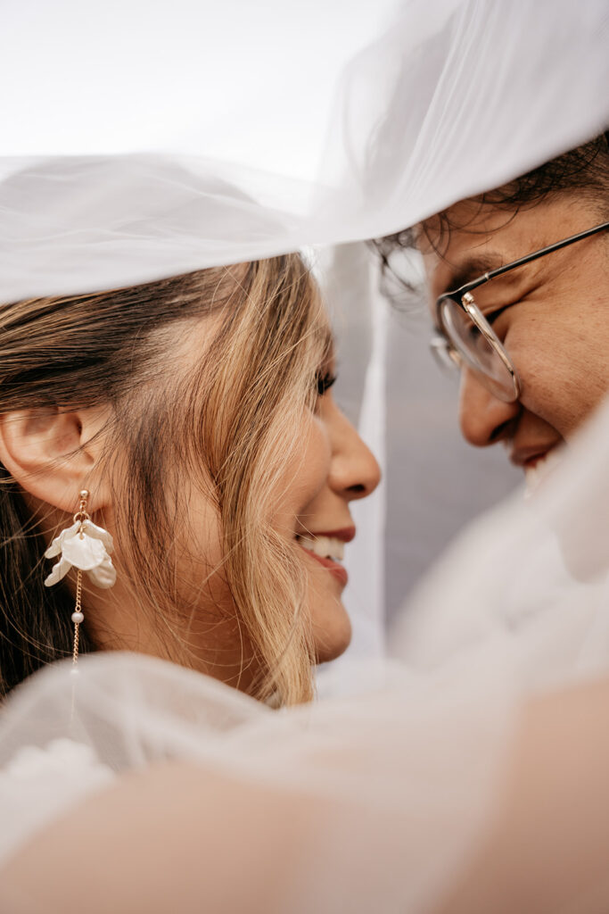 Bride and groom under veil, smiling closely.