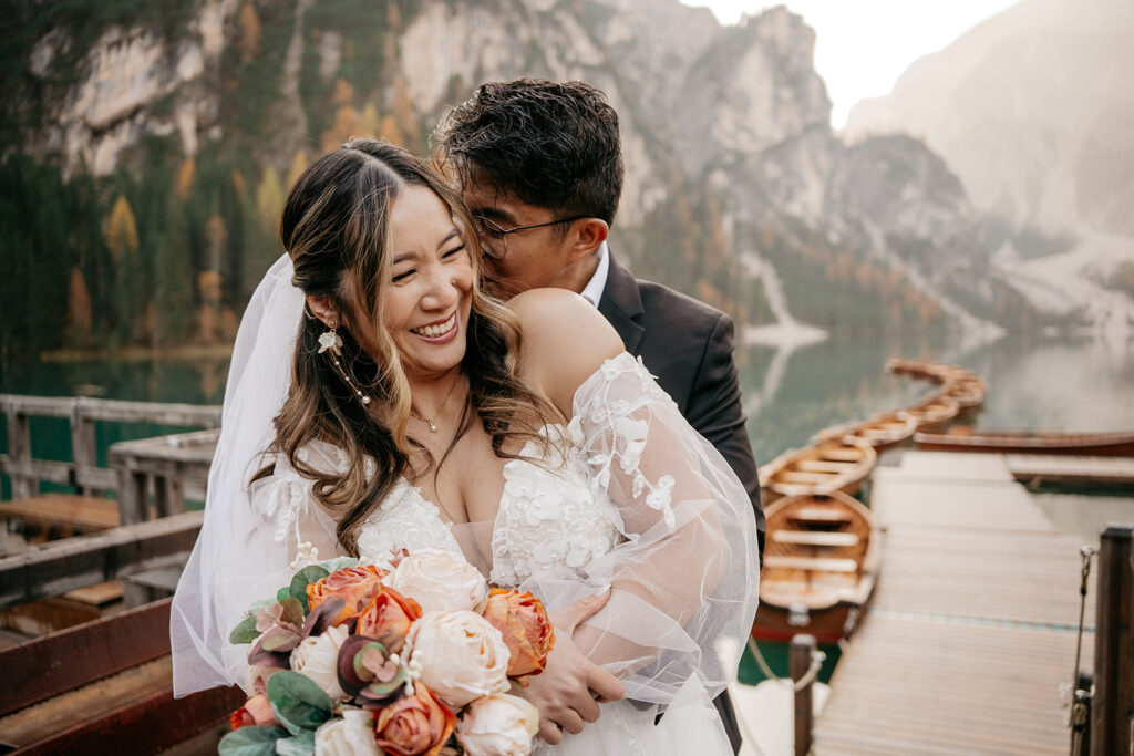 Bride and groom embrace by mountain lake.