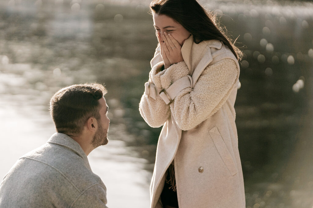 Man proposing by the water, woman surprised and happy.