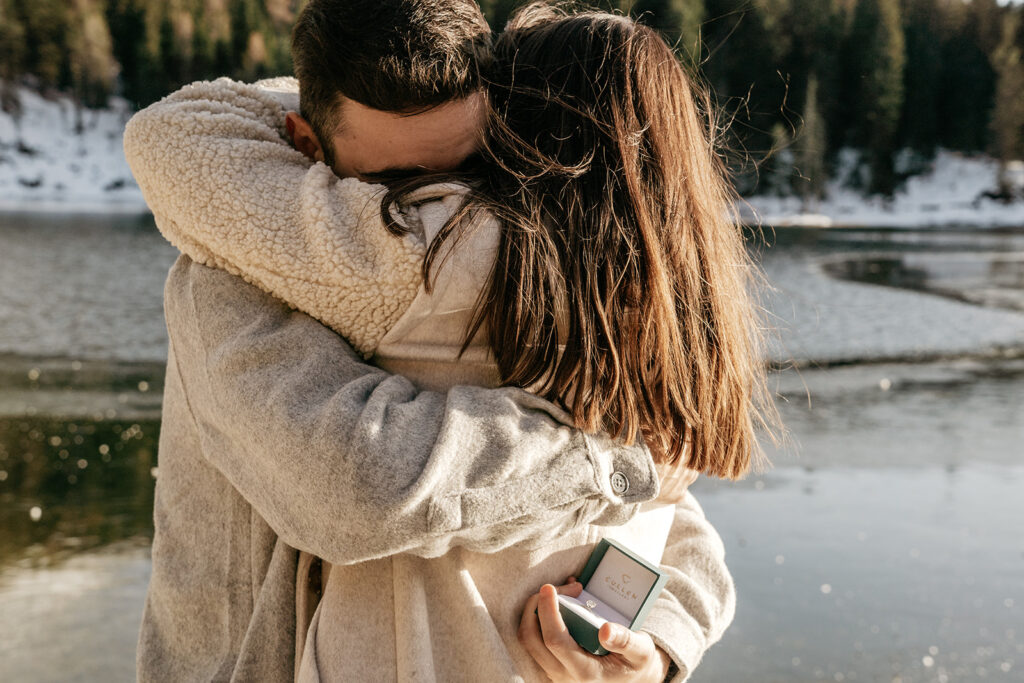 Couple hugging with engagement ring by a lake.