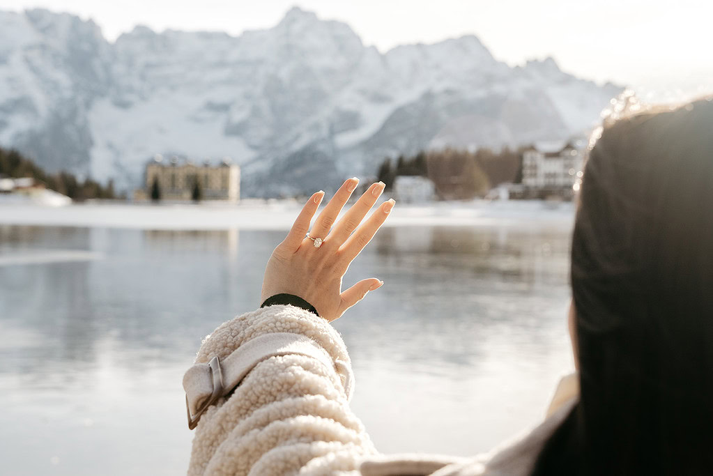 Hand with ring, snowy mountain backdrop, lake view.