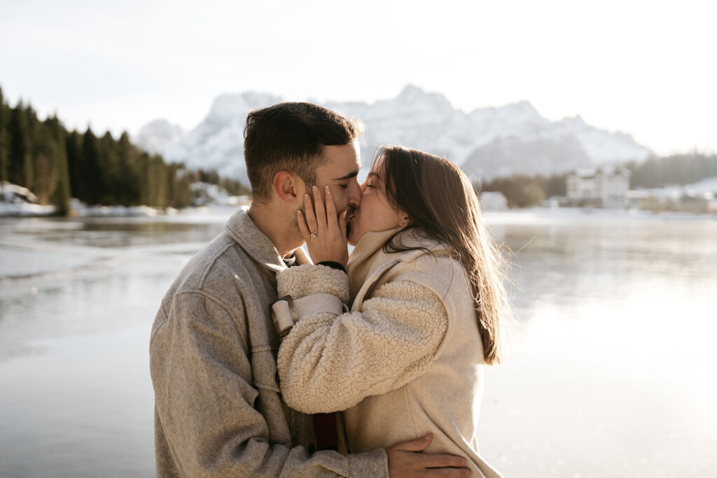 Couple kissing by snowy lake, mountains behind.