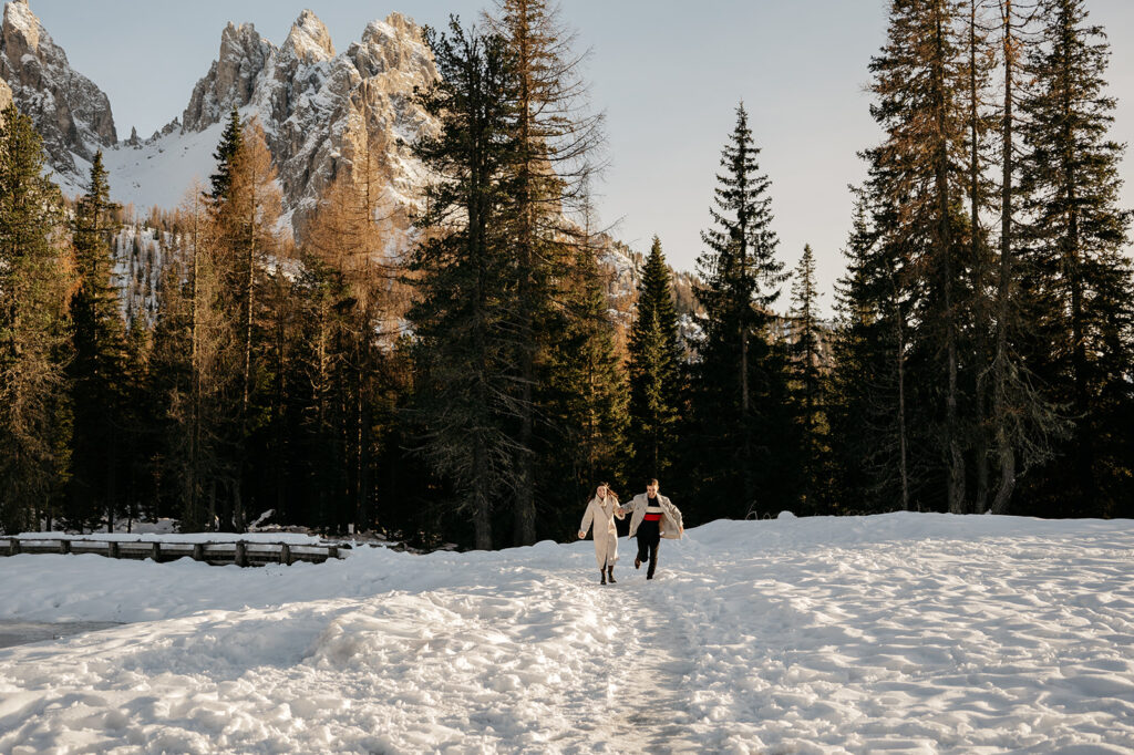 Couple walking in snowy forest with mountain backdrop.