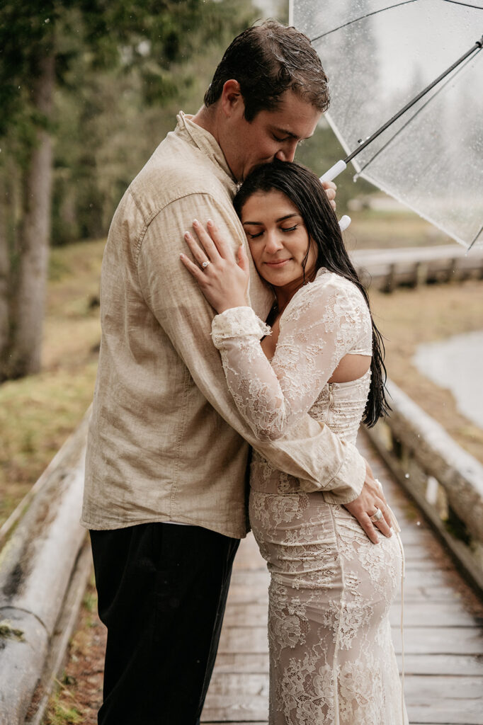 Couple embraces under umbrella in rainy forest.