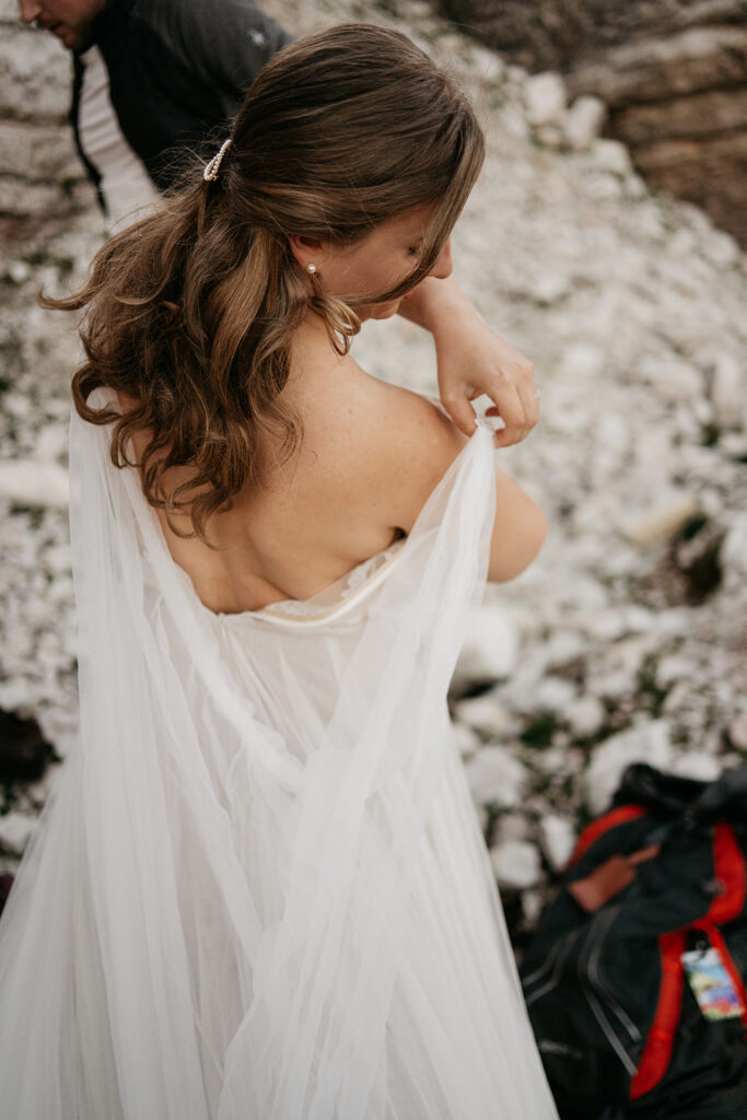 Bride adjusting white dress outdoors on rocky ground.