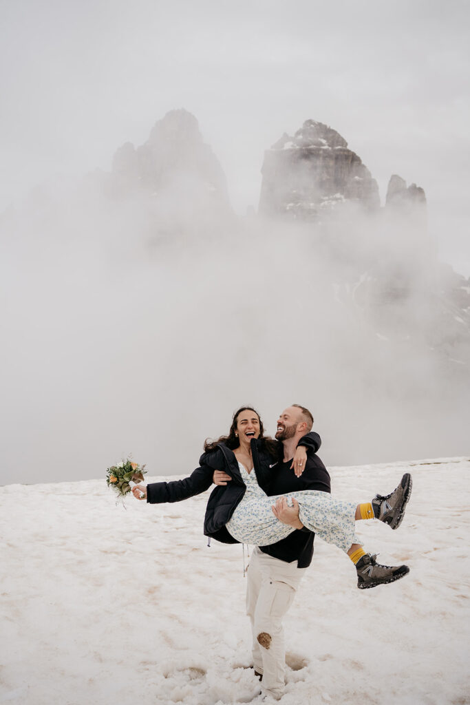 Couple laughing on snowy mountain, foggy background.