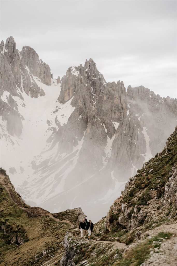 Hikers on mountain path with rocky peaks and mist.