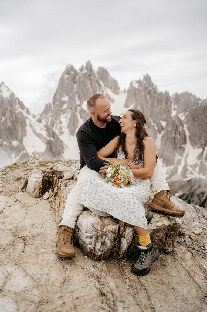 Couple sitting joyfully on mountain rock, holding flowers.