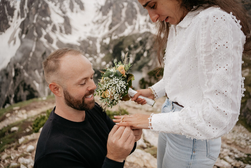 Man proposing on mountain with ring and flowers.