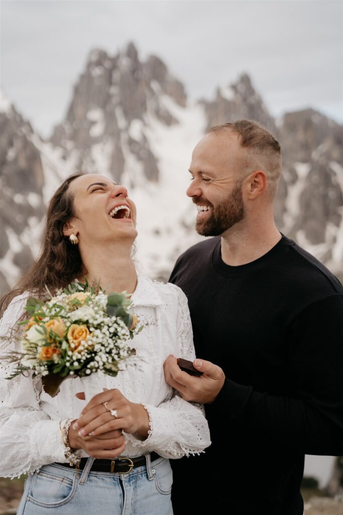 Couple laughing with bouquet, mountains in background.