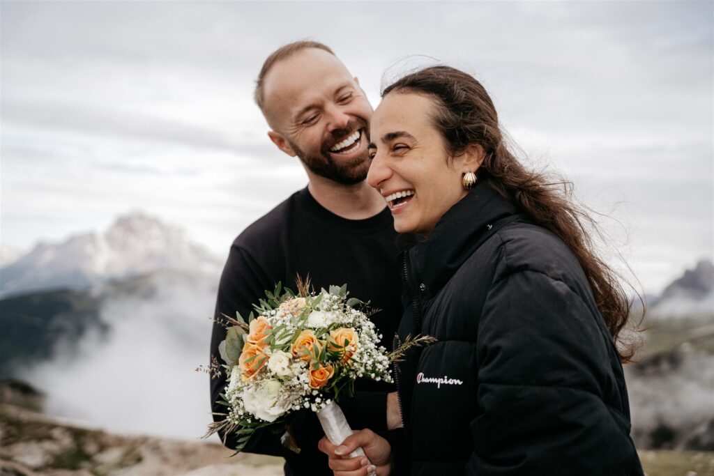 Laughing couple with bouquet in mountainous landscape.