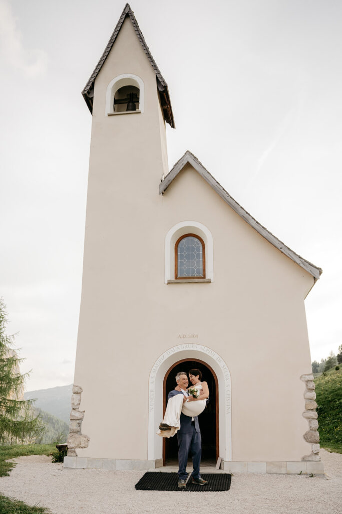 Groom carrying bride outside small chapel.