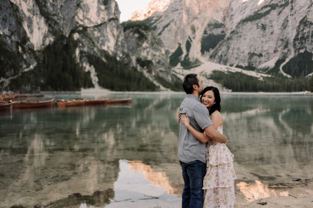 Couple hugging by mountain lake with boats.