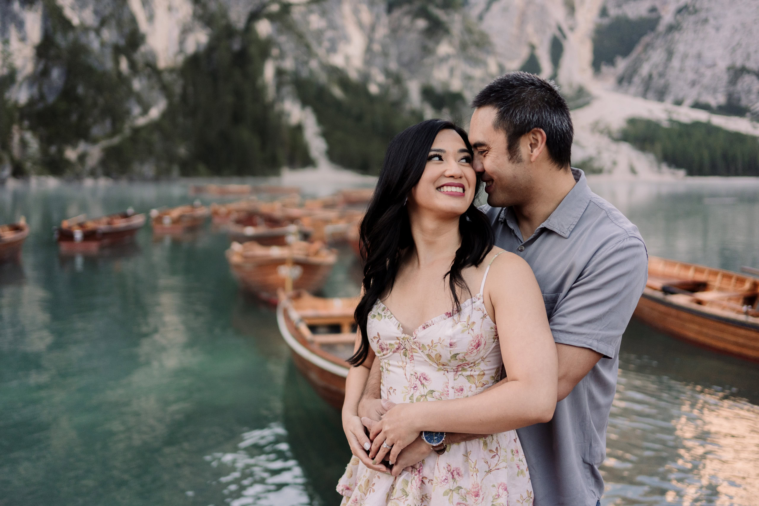 Couple embracing by scenic lake with wooden boats.