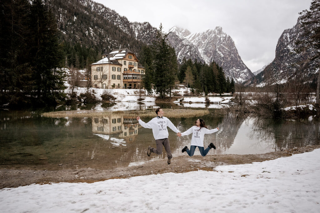 Couple jumping by snowy lake and mountain lodge.