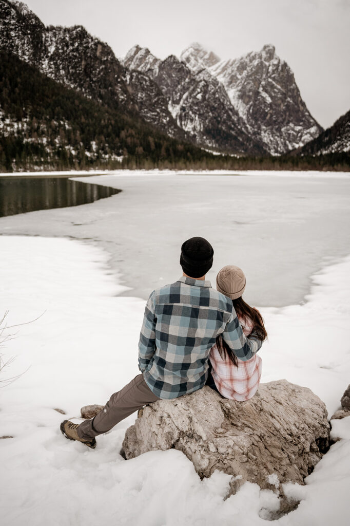 Couple sits by snow-covered mountain lake scenery.