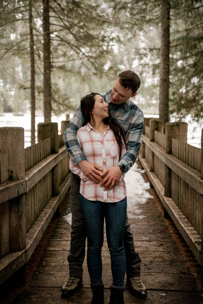 Couple embracing on snowy wooden bridge outdoors.