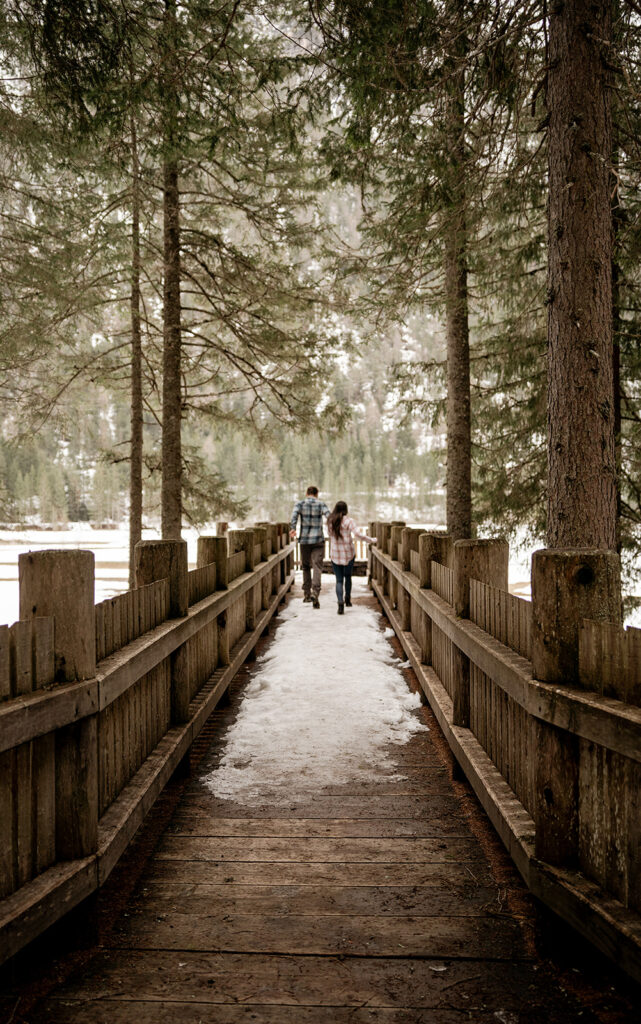 Couple walking on snow-covered bridge in forest.