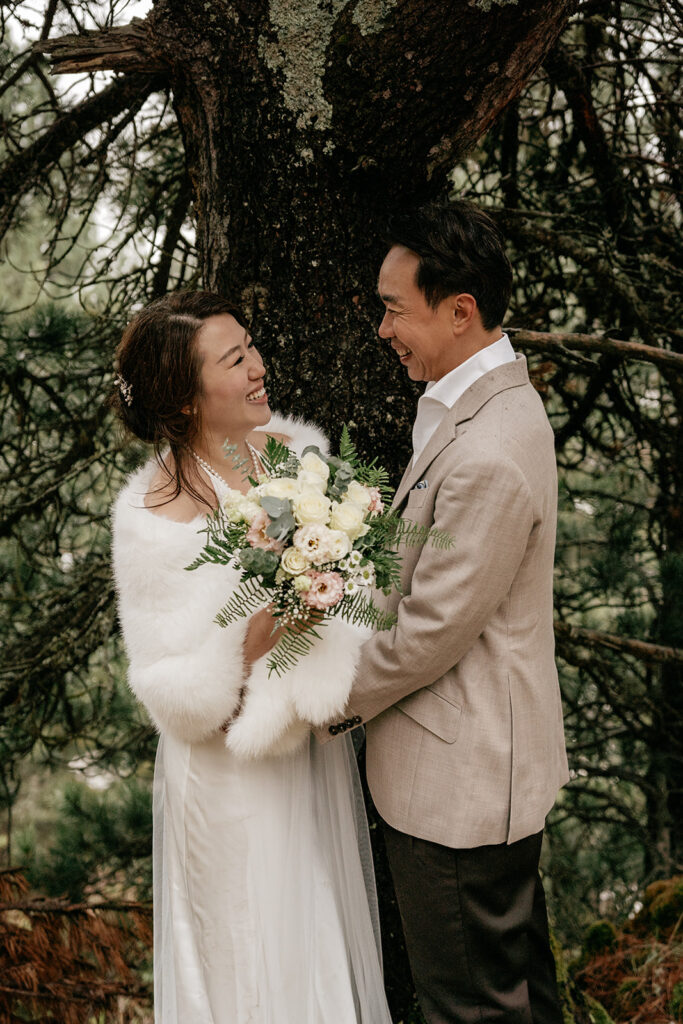 Bride and groom smiling under tree with bouquet.