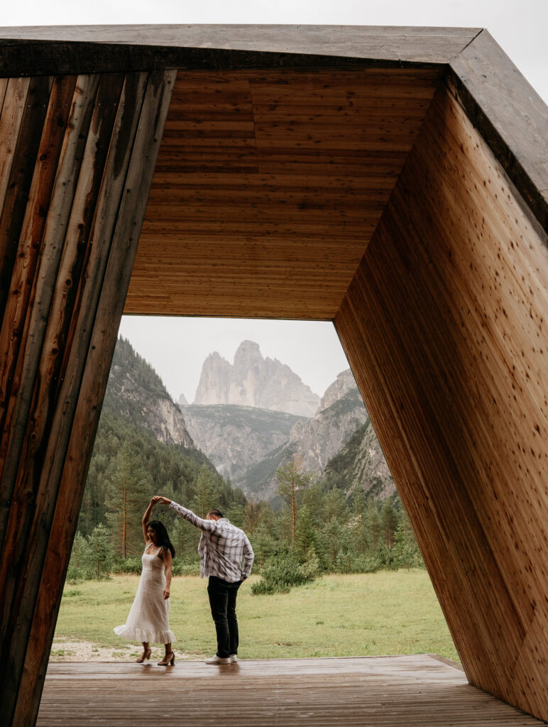 Couple dancing in mountain valley, wooden structure foreground.