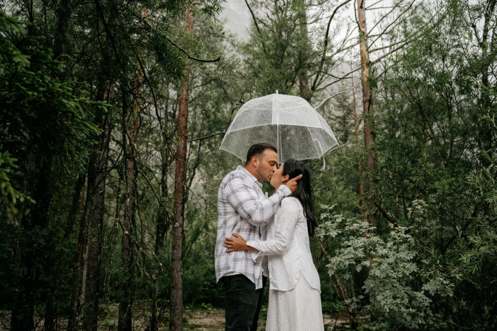 Couple kissing under umbrella in forest rain.