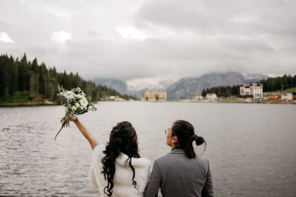 Couple by lake, bouquet raised, mountainous backdrop