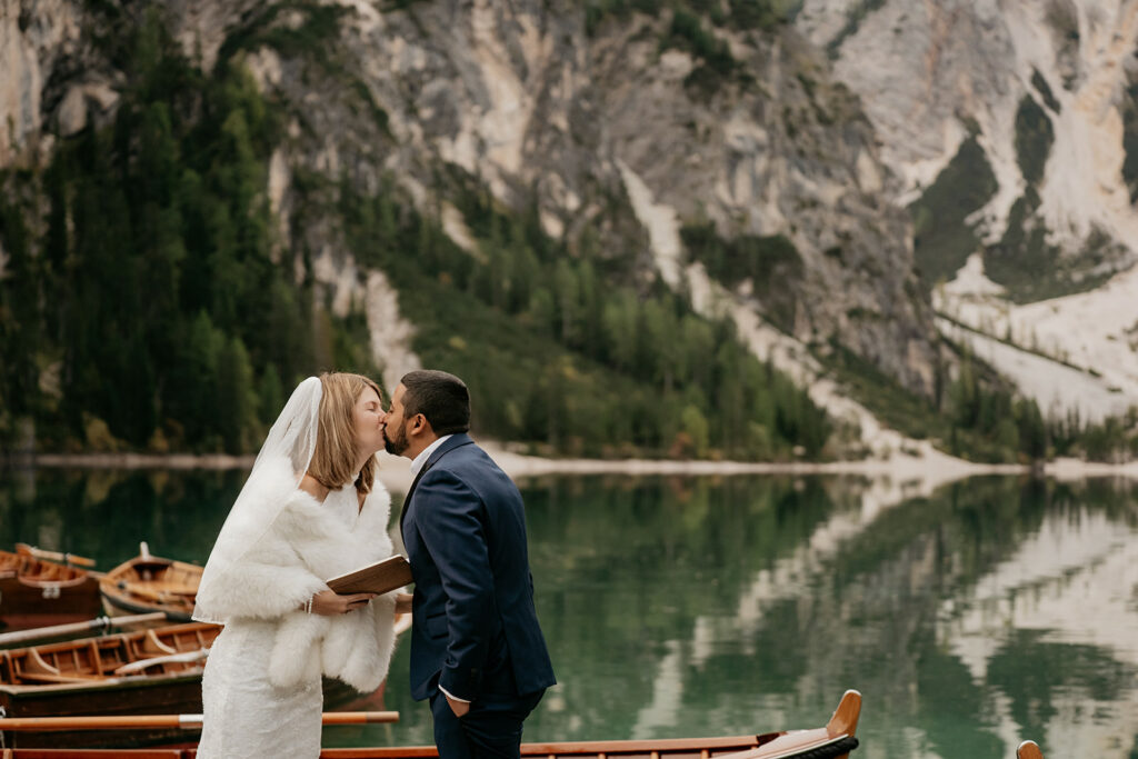 Couple kissing by mountain lake with boats.