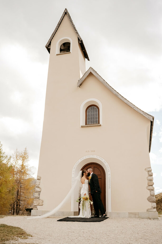 Couple kissing in front of a rustic chapel.