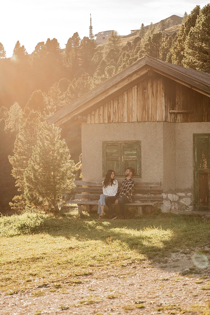 Couple sitting on bench near mountain cabin at sunset.
