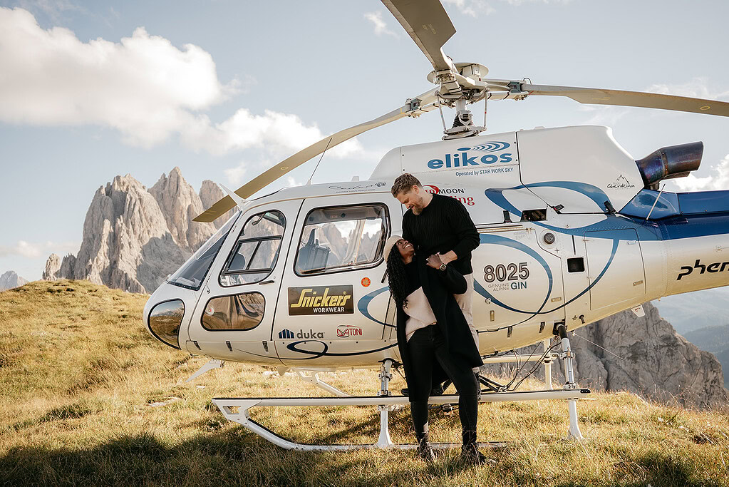 Couple embracing near helicopter, mountains in background.