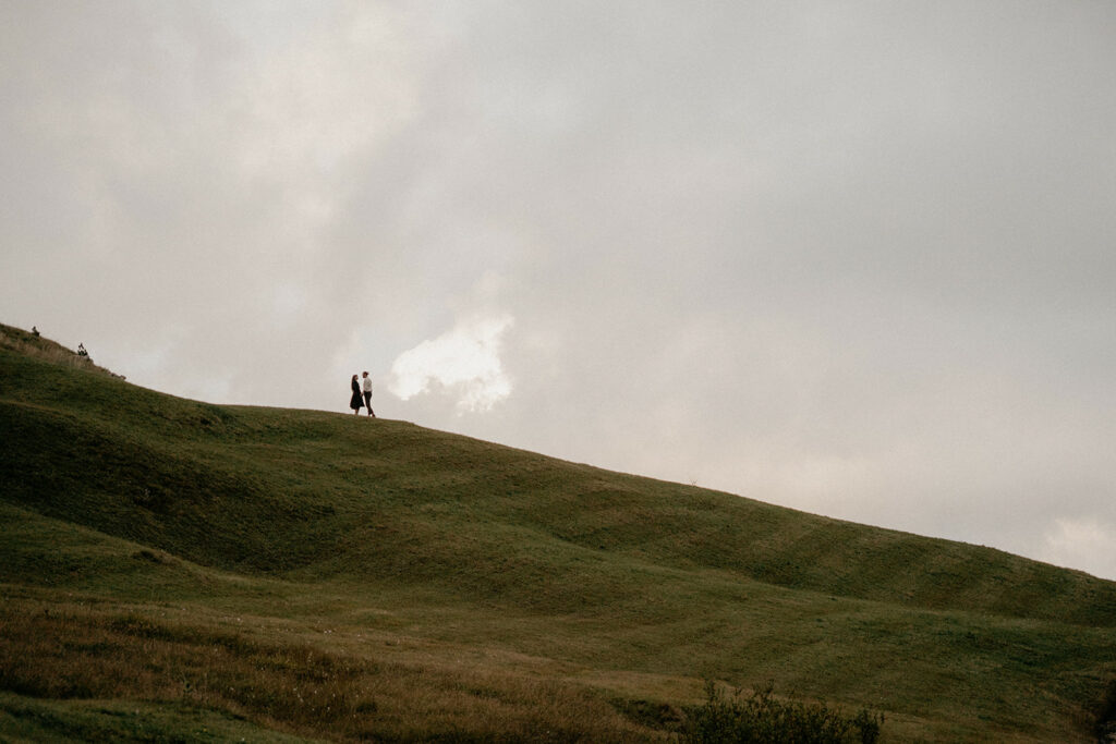Couple walking on grassy hill under cloudy sky.