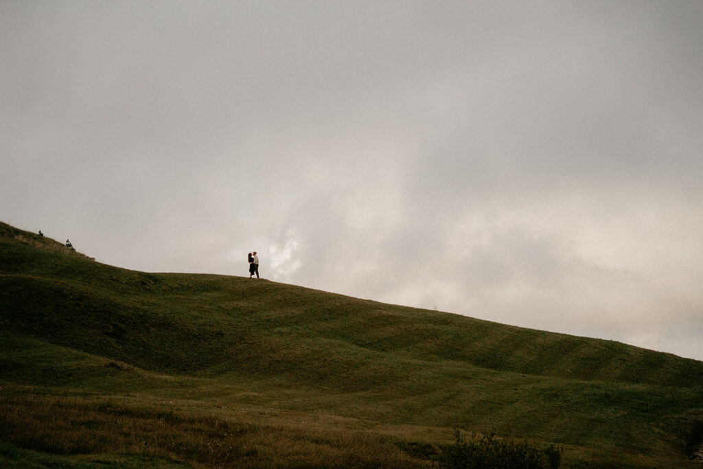 Couple standing on grassy hill under cloudy sky.