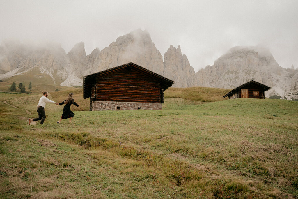 Couple running near mountain cabins under cloudy sky.