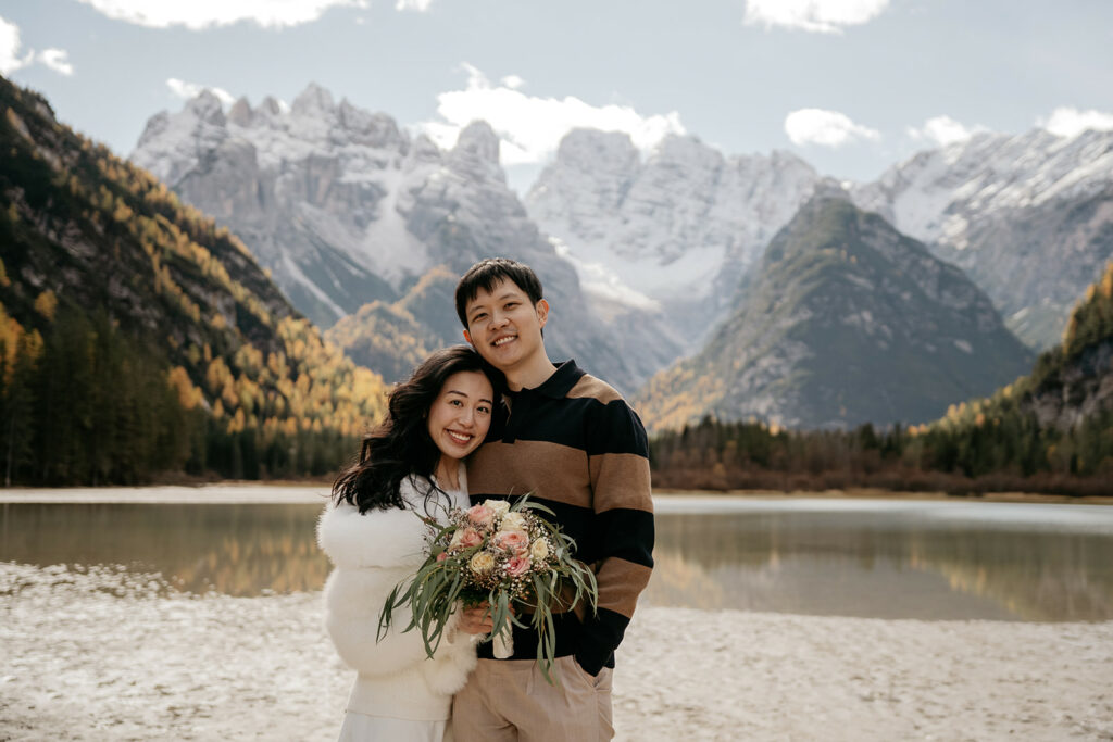 Couple smiling by mountain lake with bouquet.